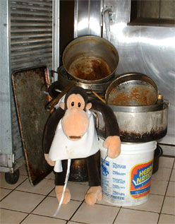 Monkey stands before a pile of dirty pots/pans taller than he.
